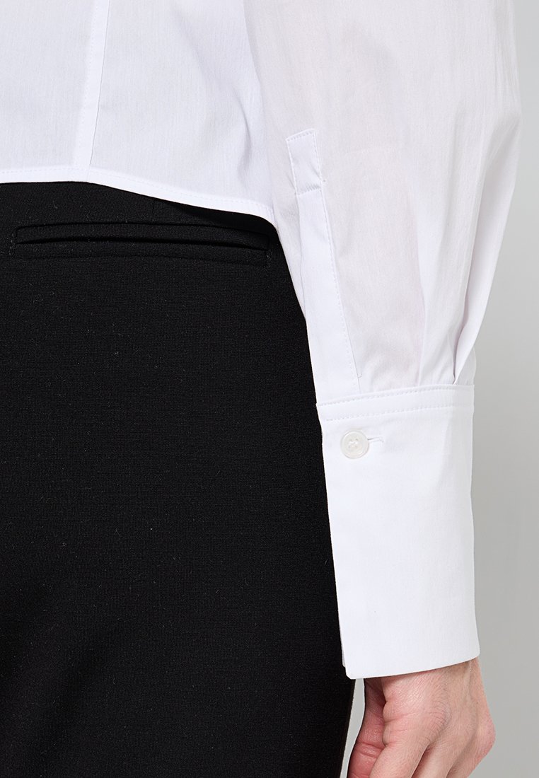 Weißes Baumwollhemd mit aufgeschlagenem Ärmel und einem einzelnen Knopfverschluss. Kombiniert mit schwarzen, maßgeschneiderten Hosen und einem eleganten Taschendesign.