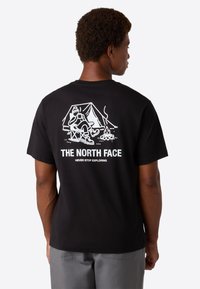 Mand iført sort T-shirt med hvid campinggrafik, der viser en person, der slapper af ved telt og lejrbål, samt teksten "THE NORTH FACE".