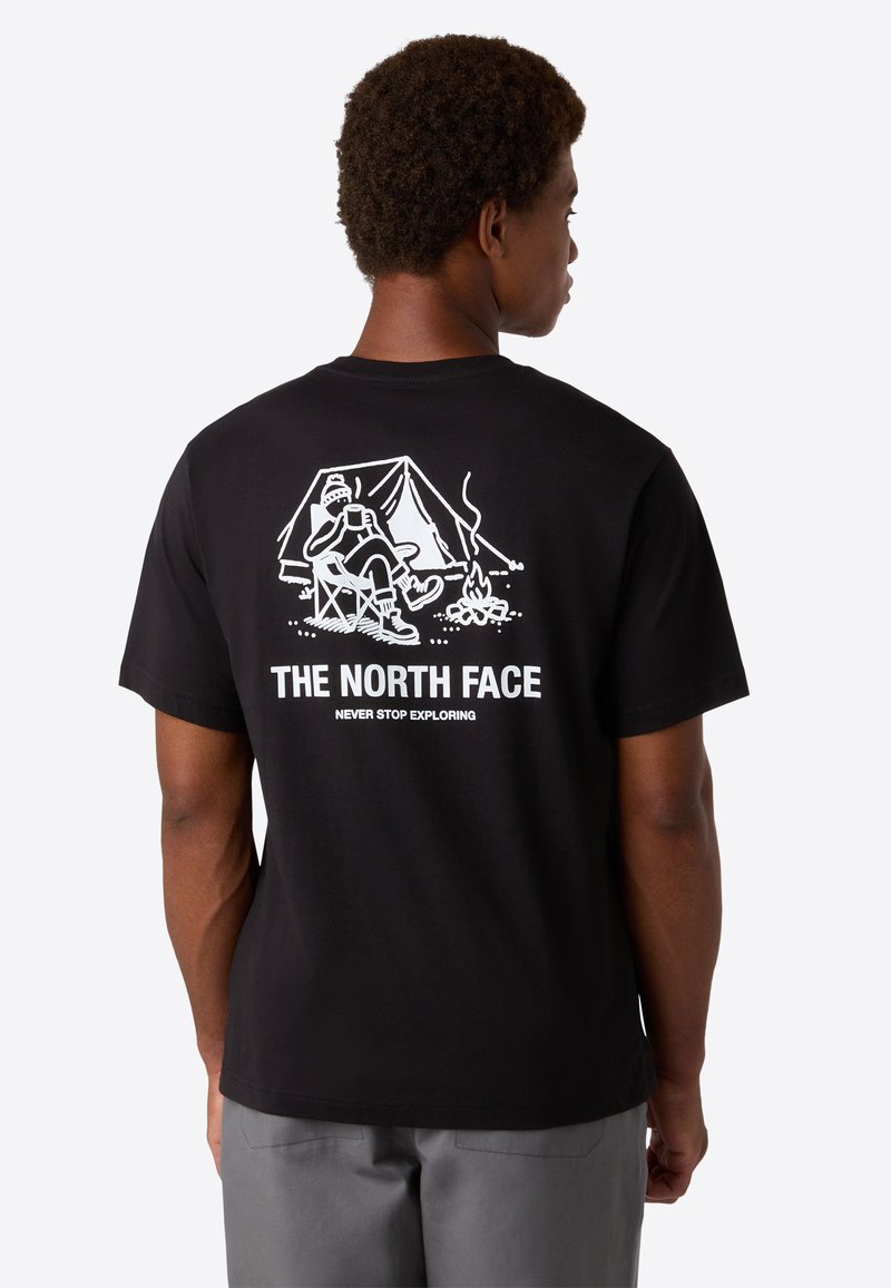 Mand iført sort T-shirt med hvid campinggrafik, der viser en person, der slapper af ved telt og lejrbål, samt teksten "THE NORTH FACE".