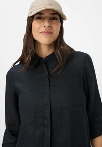 Schwarzes Leinen-Hemd mit Knöpfen, Kragen und aufgerollten Ärmeln, getragen mit einer beigen Baseballkappe. Glatte Textur, minimalistisches Design.