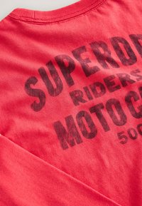 Rød bomulds langærmet skjorte med sort grafisk tekst, der lyder "SUPERDRY RIDERS MOTO CO 500" på forsiden. Blød tekstur, afslappet design.