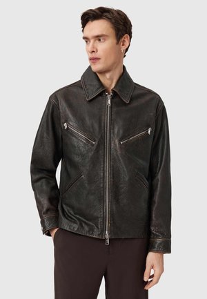 Brązowa skórzana kurtka o gładkiej fakturze, z ostrym kołnierzem i trzema kieszeniami na zamek. Posiada przedni zamek błyskawiczny oraz przeszycia dekoracyjne.