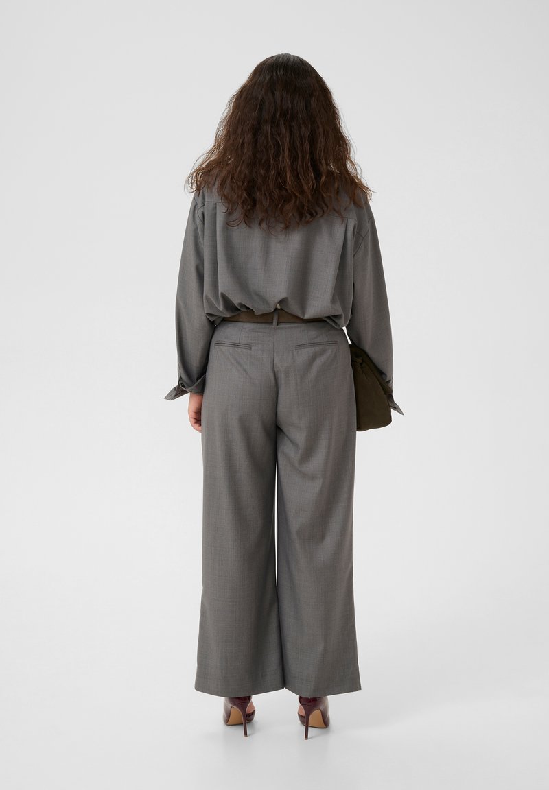 Grau karierte Bluse und weite Hosen aus strukturiertem Stoff. Das Outfit wird durch eine dunkelgrüne Tasche und braune High Heels ergänzt.