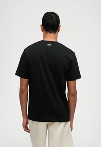 Μαύρη βαμβακερή μπλούζα με κοντά μανίκια, κλειστή λαιμόκοψη και πράσινο κεντητό λογότυπο στον αυχένα. Λείος υφασμάτινος σχέδιο, κανονική εφαρμογή.