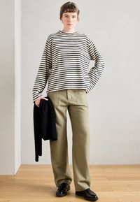 Camisola de manga longa riscada em bege e preto, combinada com calças largas verde-azeitona. Segurando um suéter de malha preto dobrado, usando sapatos pretos.