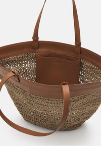 Sac en paille tissée couleur fauve avec garniture et poignées en cuir marron, doté d'une poche intérieure discrète. Forme ronde, corps texturé, design minimaliste.