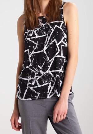 Femme portant un haut noir sans manches avec des formes géométriques abstraites blanches, assorti à un pantalon gris, sur un fond uni.