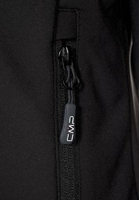 Nahaufnahme eines Reißverschlusses einer schwarzen Jacke mit einer Textil-Ziehlasche, die mit "CMP" beschriftet ist, vor einem schwarzen strukturierten Stoff.
