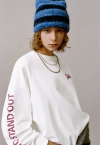 Felpa bianca oversize con la scritta "STAND OUT" in bordeaux sulla manica, catene d'argento sovrapposte e un cappello a maglia a righe blu.