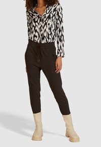 Sort-hvid mønstret skjorte med en lomme, parret med brune jogger-stil bukser med elastisk talje og snørrebånd, samt cremefarvede støvler.