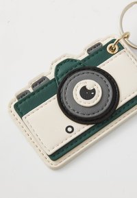 Green and cream rubber keychain shaped like a camera, featuring a prominent black lens detail with gray accents and stitched edges.