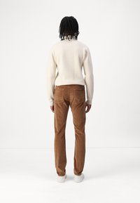 Pull en tricot beige, pantalon en velours côtelé marron, baskets blanches à tige basse, montrés de dos sur fond uni.
