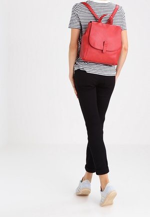 Henkilö, jolla on mustat housut, raidallinen paita, valkoiset lenkkarit ja punainen nahkainen reppu, kävelee yksiväristä valkoista taustaa vasten.