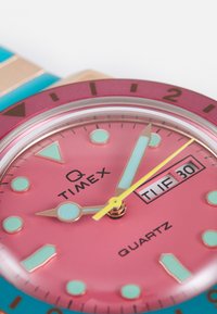 Timex Q kellal on roosa sihver, kuldne riistvara, türkiissinised aktsendid, kuupäevanäidik ja mitmevärvilised tunni märgid koos kvartsliikumisega.