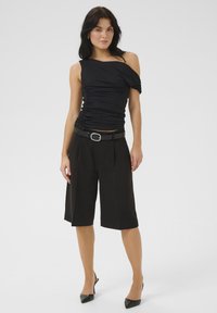 Zwarte gerimpelde top met een asymmetrische halslijn, gecombineerd met high-waisted zwarte culottes, versterkt door een zwarte riem en hoge schoenen.