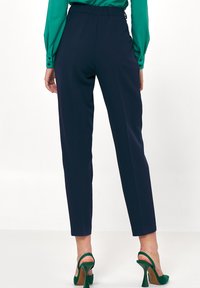 Spodnie w marynarskim granacie o wysokim stanie i prostym kroju nogawki. Gładka tkanina i subtelne przednie kanty. Połączone z zielonymi sandałami na obcasie.