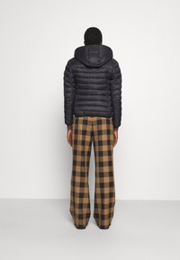 Doudoune bleu marine foncé avec des coutures verticales, une capuche et des poignets élastiques, associée à un pantalon à jambes larges à carreaux marron et noir.