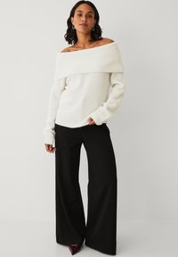 Chunky hvid off-the-shoulder sweater med ribbet tekstur, parret med høj-stigede sorte brede bukser og burgundy hæle.