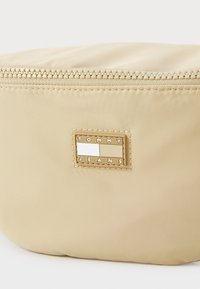 Borsa cosmetica in nylon beige con chiusura a zip e un'etichetta rettangolare del marchio con toni bianchi e beige. Texture liscia, forma arrotondata.