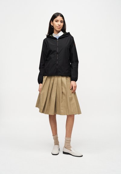 Femme portant une veste à capuche noire zippée, une jupe midi plissée beige, des chaussettes beiges et des mocassins blancs, debout devant un fond blanc uni.