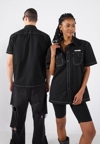Magliette nere a maniche corte con cucitura bianca, doppie tasche sul petto e bottone frontale, indossate con pantaloni neri strappati e pantaloni da bici.