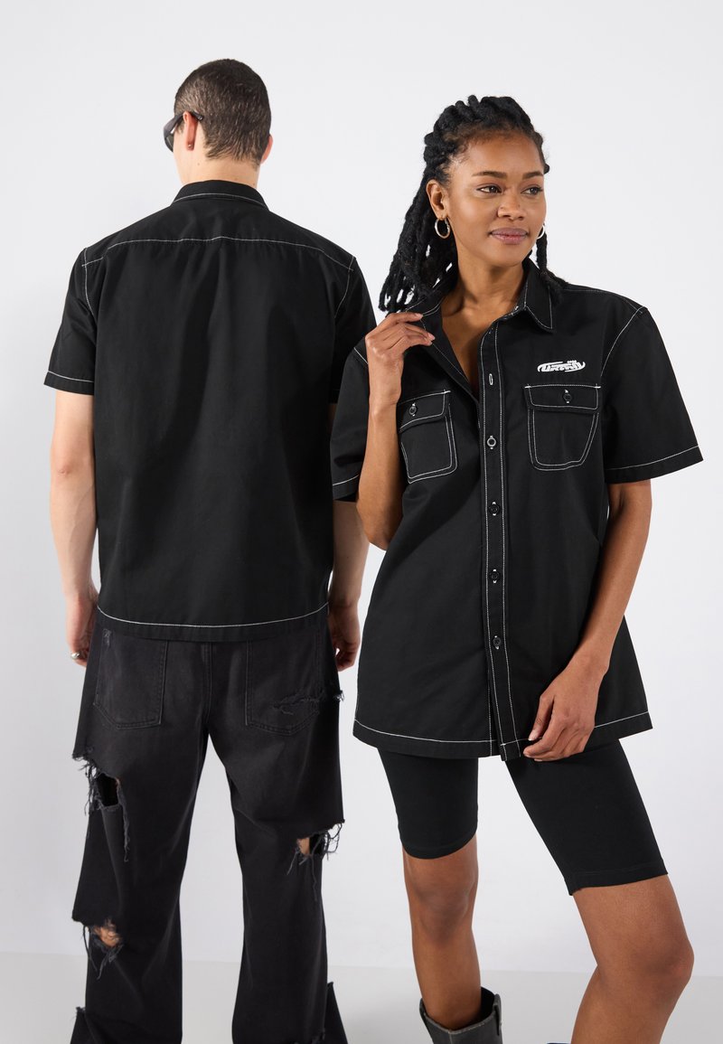 Magliette nere a maniche corte con cucitura bianca, doppie tasche sul petto e bottone frontale, indossate con pantaloni neri strappati e pantaloni da bici.