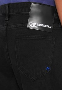 Črne denim hlače z sijočim črnim usnjenim žepom, ki nosi napis "KARL LAGERFELD JEANS" v beli barvi. Modri "K" akcent na zadnjem žepu.