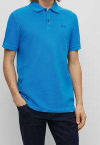 Blå pikétröja med krage, korta ärmar och två-knappsstängning. Har en subtil logotyp på bröstet och kombineras med mörka jeans.