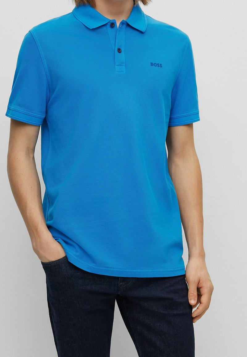 Blå pikétröja med krage, korta ärmar och två-knappsstängning. Har en subtil logotyp på bröstet och kombineras med mörka jeans.