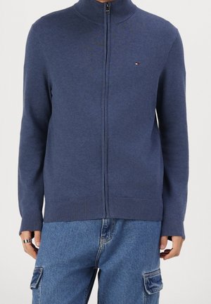 Person iført en blå lynlås-sweater med lille logo på brystet og blå denim cargo bukser med lommer i siderne.