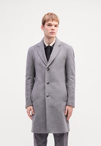 Cappotto di lana grigio con un design su misura, cinque bottoni e rever appuntiti. Indossato sopra una camicia scura con colletto chiaro.