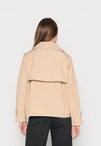 Beige trench jacket with a wide collar, oversized fit, layered back design, and buttoned cuffs, paired with dark jeans.