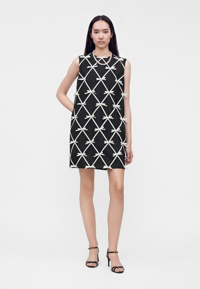 Femme debout portant une robe noire sans manches avec des motifs de nœuds blancs, un collier de perles et des sandales à talons hauts noires sur un fond uni.