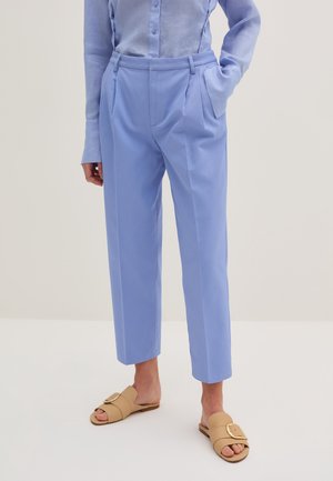 Person trägt hellblaue, maßgeschneiderte Hose mit Falten, ein passendes blaues Hemd zum Knöpfen und beigefarbene Slide-Sandalen mit großen Schnallen.