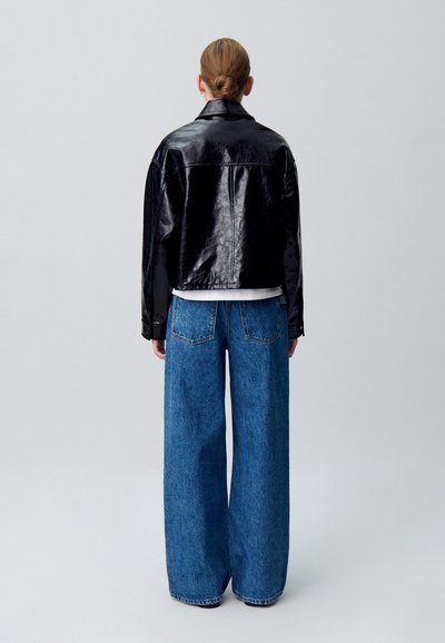 Giacca di pelle nera cropped con texture lucida abbinata a jeans in denim blu a gamba larga. Sfondo grigio uniforme.