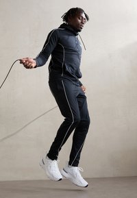 Juoda sportinė treniruočių apranga su baltais šonais ir užtrauktuku, derinama su baltais sportbačiais; asmuo šoka su šokinėjimo virve.