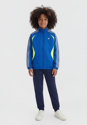 Otrok z kodrastimi lasmi, oblečen v modro in neonsko rumeno Adidas trenirko jakno, temno modre hlače in bele superge, stoji in se smehlja.