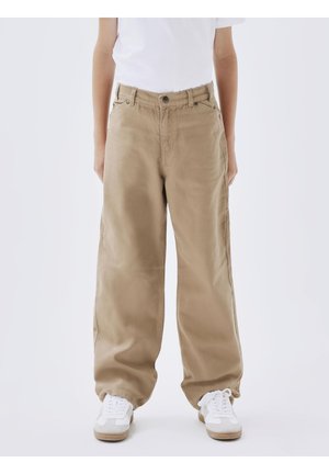 Persoon die loszittende beige broek, witte sneakers en een wit shirt draagt, staand tegen een effen lichte achtergrond.