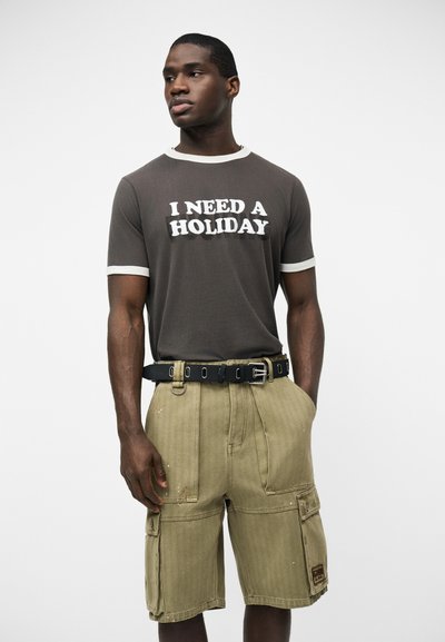 Νέος άνδρας φοράει σκουρόχρωμο μπλουζάκι με το κείμενο «ΧΡΕΙΑΖΟΜΑΙ ΔΙΑΚΟΠΕΣ» και μπεζ cargo σορτς, στέκεται με τα χέρια στις τσέπες μπροστά σε λευκό φόντο.
