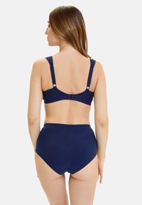 Femme aux longs cheveux bruns portant un soutien-gorge bleu marine et un slip taille haute, vue de dos sur un fond blanc uni.