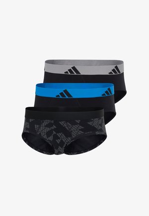 Athletischer Mann steht barfuß, trägt schwarze Slips mit einem blauen Bund und einem schwarzen Adidas-Logo, blickt nach rechts vor einem schlichten weißen Hintergrund.