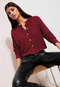 Cardigan burgundiu texturat, cu gât rotund și nasturi aurii, purtat peste pantaloni negri din piele. Design neted, mulat, cu mâneci lungi.