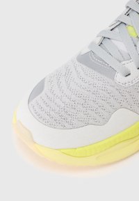 Punta di sneaker grigio chiaro con tomaia in maglia a motivo, lacci grigi chiari, sovrapposizione in suede e suola gialla imbottita su uno sfondo bianco.