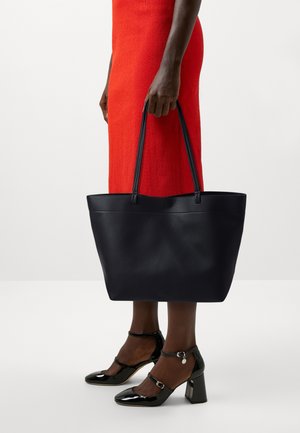 Grande borsa tote in pelle nera con superficie texturizzata, manici allungati e design minimalista. Indossata insieme a scarpe con tacco in vernice nera.