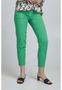 Green slim-fit trousers made of smooth fabric, featuring a sleek design, ankle length, and paired with low-heeled black slide sandals.
