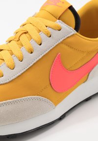 Gul sportssko med en texturerad mocka- och andningsbar tygövre, rosa Nike-logga, vadderad krage och svart sula.