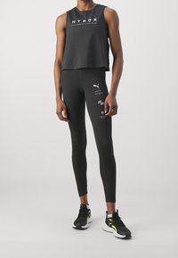 Top corto nero con testo bianco "HYROX", abbinato a leggings neri aderenti con accenti di logo bianchi e sneakers nere con dettagli neon.