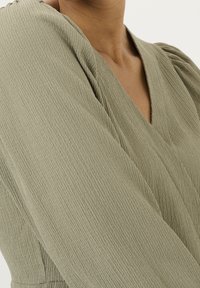 Olivgrünes, strukturiertes Kleid mit V-Ausschnitt und Puffärmeln, das dezente Rippdetails im Stoff zeigt.