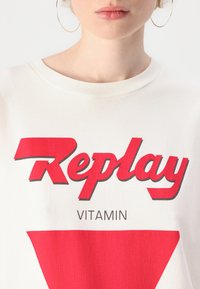 Biała bawełniana koszulka z krótkim rękawem, z wyraźnym czerwonym napisem "Replay" oraz graficznym wzorem trójkąta. Okrągły dekolt, gładka tekstura.