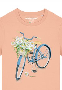 Perzikkleurig t-shirt met een blauwe fietsafbeelding, waarop een gevlochten mand is te zien die gevuld is met witte madeliefjes en groene bladeren; gedetailleerd ontwerp van het wiel en de pedaal.